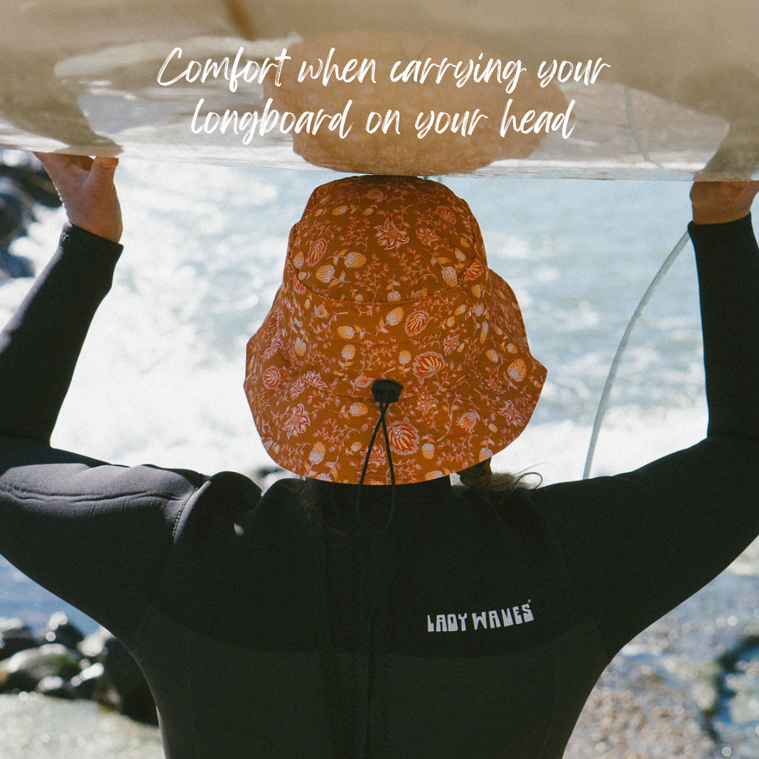 Woman wearing patterned surf hat surfshell best protection out surfing waves. carrying a longboard surfboard on her head with ease due to added comfort from the helmet