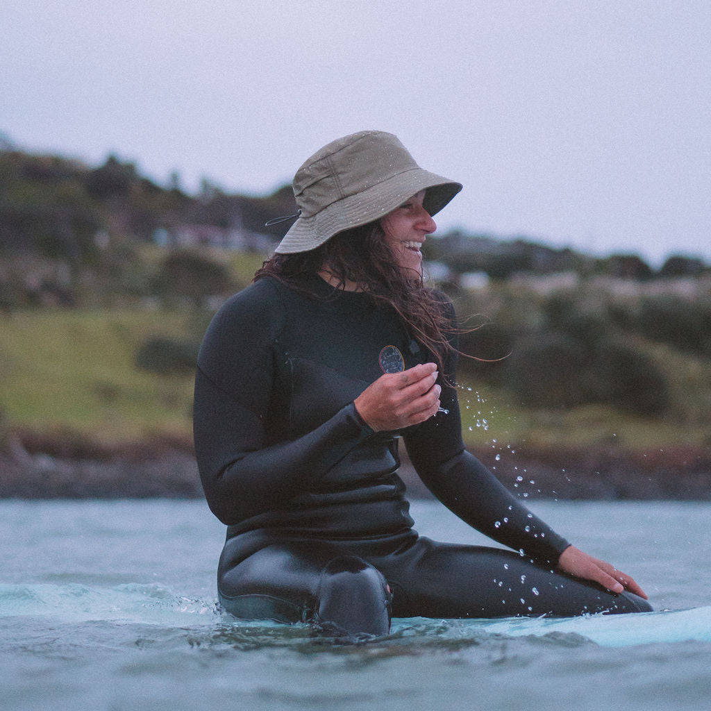 Person in a wetsuit and hat sitting on a surfboard in water with a scenic background