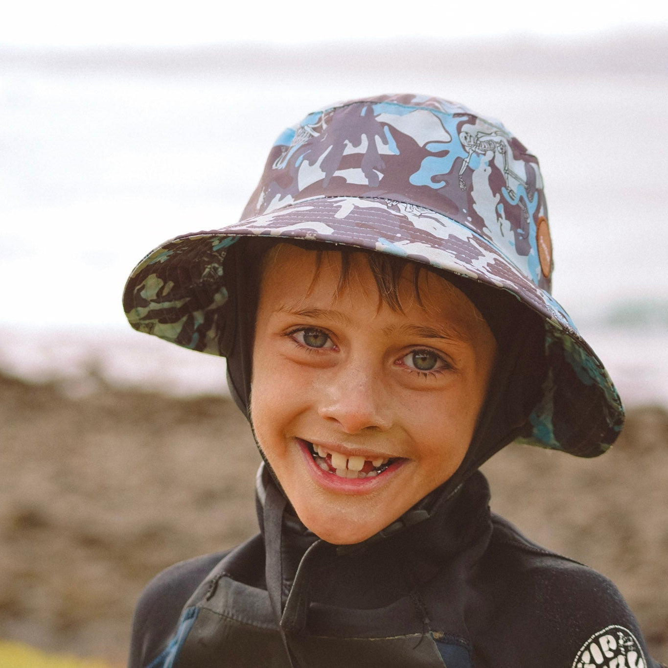 Child wearing a patterned hat and wetsuit by a body of water