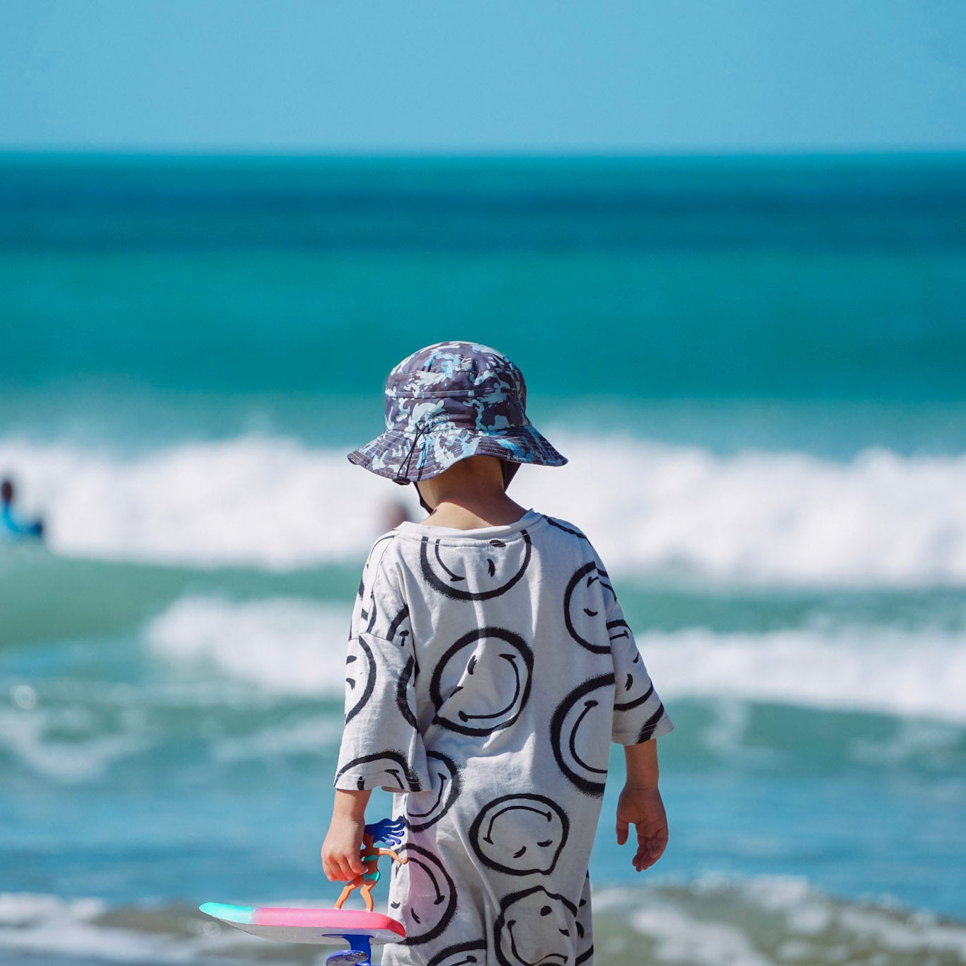 Child in a sun hat and swimsuit standing in shallow water at the beach with ocean waves in the background.