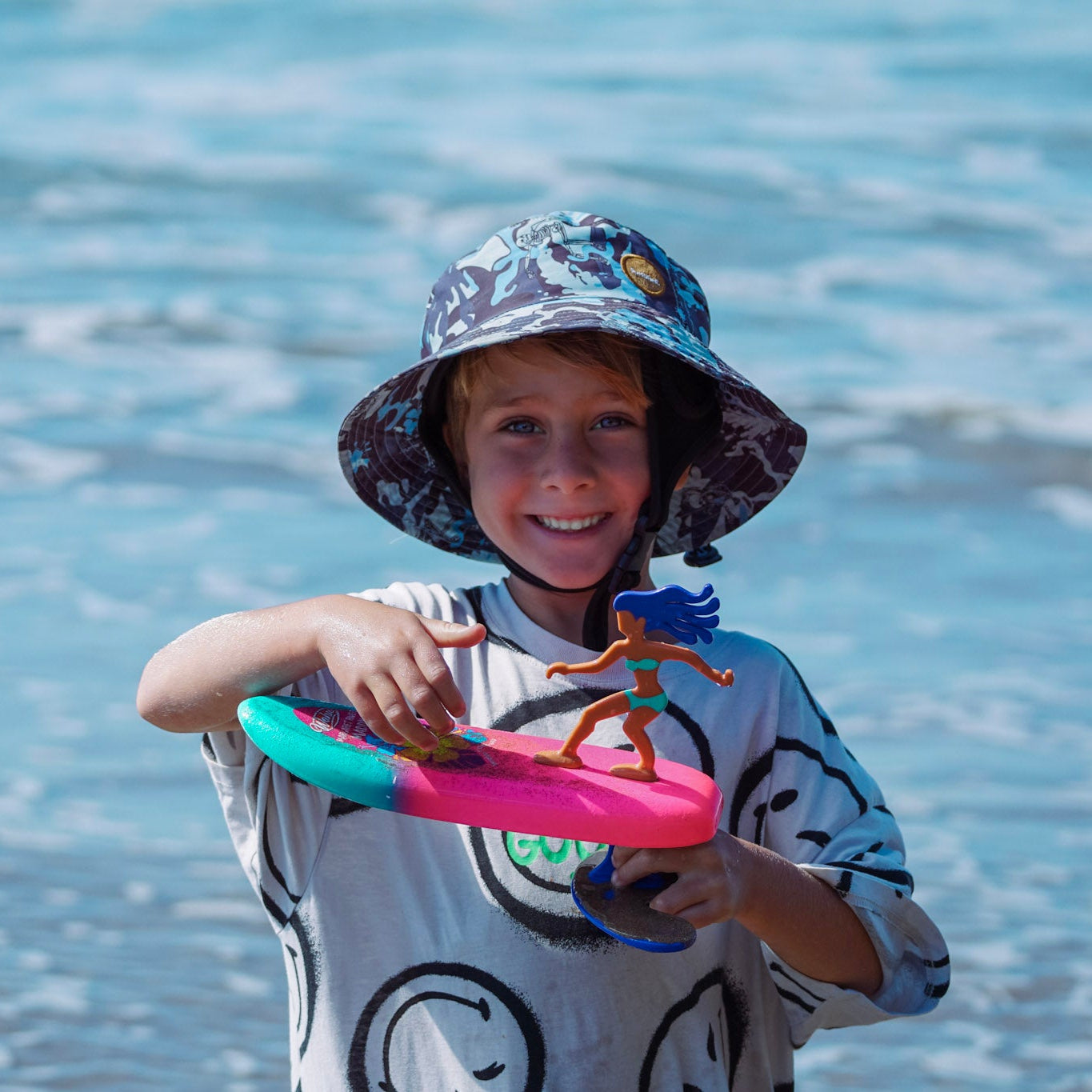 Child holding a toy in front of a body of water wearing a surfshell protective surf hat