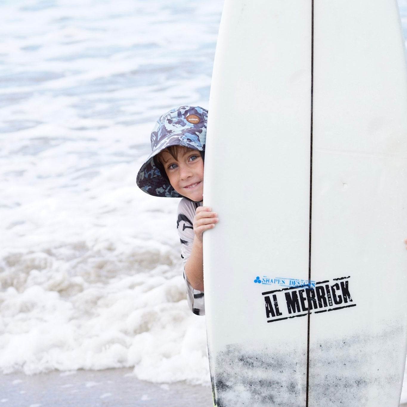 Child peeking out from behind a surfboard with Al Merrick logo at the beach, the child is wearing a surfshell protective surf hat blue camo colour with surfing skeletons 