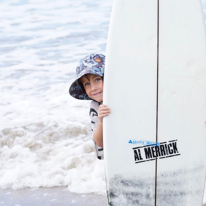 Child peeking out from behind a surfboard with Al Merrick logo at the beach, the child is wearing a surfshell protective surf hat blue camo colour with surfing skeletons 
